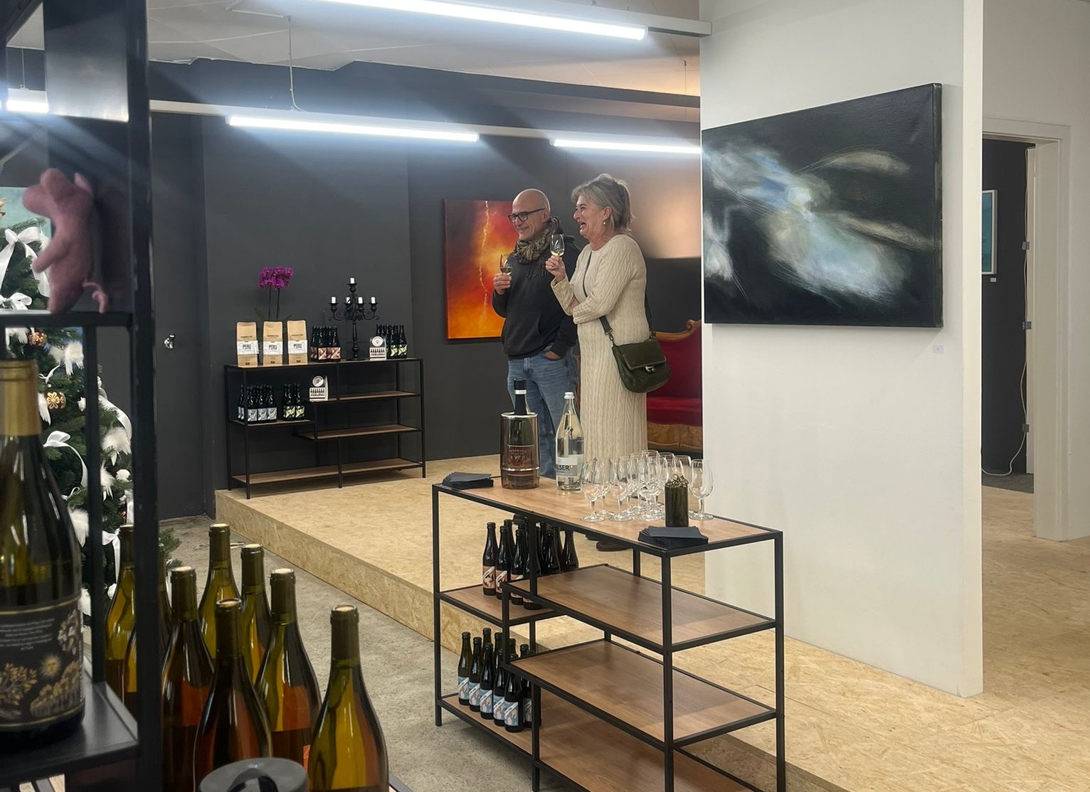 Ausstellungsansicht im Kitchen Angel, Dezember 2025: Besucherinnen und Besucher mit Glas vor den ausgestellten Bildern