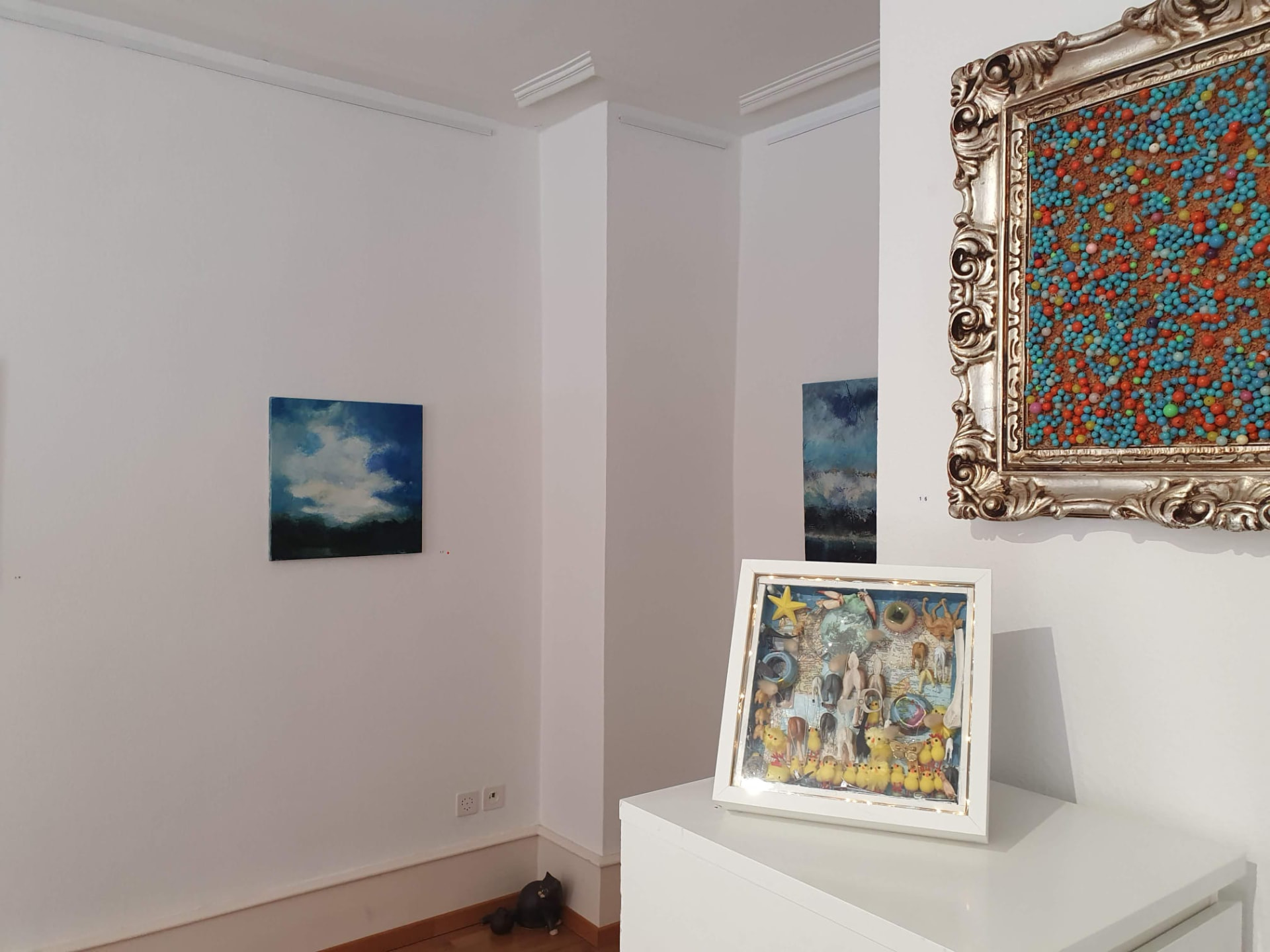 Ausstellungsansicht Kunstspeicher Wiederkehr: beleuchtete Objektbox auf weissem Schrank, darüber weitere Bilder